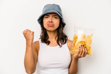 Beyaz arka planda izole edilmiş bir paket cipsi tutan genç İspanyol kadın yumruğunu kameraya gösteriyor, agresif bir yüz ifadesi..