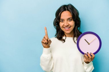 Mavi arka planda bir saat tutan genç İspanyol kadın bir numarayı parmağıyla gösteriyor..