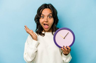 Mavi arka planda bir saat tutan genç İspanyol kadın şaşırdı ve şok oldu..