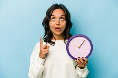 Elinde mavi arka planda izole edilmiş bir saat tutan genç İspanyol kadın ağzı açık bir şekilde üst tarafı işaret ediyor..