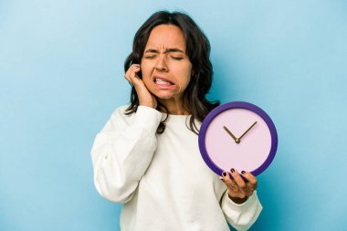Mavi arka planda bir saat tutan genç İspanyol kadın kulaklarını elleriyle kapatıyor..