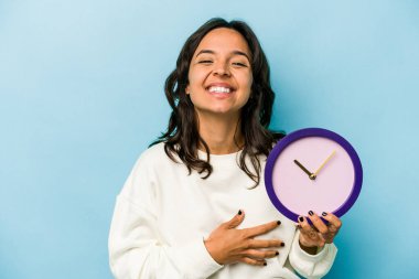 Mavi arka planda bir saat tutan genç İspanyol kadın gülüyor ve eğleniyor..