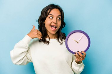 Mavi arka planda bir saat tutan genç İspanyol kadın parmaklarıyla bir cep telefonu hareketi gösteriyor..