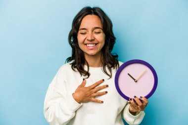 Mavi arka planda izole edilmiş bir saati tutan genç İspanyol kadın elini göğsünde tutarak kahkahalar atıyor..