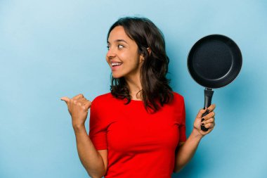 Genç İspanyol kadın elinde bir tava tutuyor mavi arka planda izole edilmiş başparmak uzakta, gülüyor ve kaygısız.
