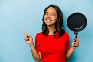 Mavi arka planda izole edilmiş bir tavayı tutan genç İspanyol kadın gülümsüyor ve boş alanda bir şeyler gösteriyor..