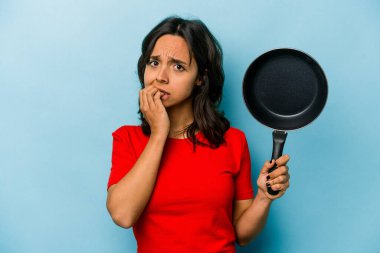 Genç İspanyol kadın elinde bir tava tutuyor mavi arka planda izole edilmiş tırnak yiyor, gergin ve endişeli..