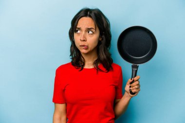 Mavi arka planda izole edilmiş bir tavayı tutan genç İspanyol kadın şaşkın ve kararsız hissediyor..