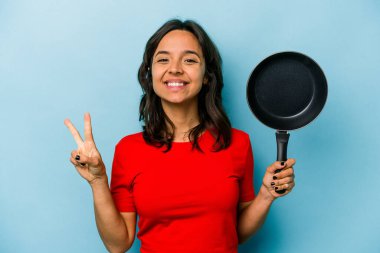 Mavi arka planda izole edilmiş tavayı tutan genç İspanyol kadın iki numarayı parmaklarıyla gösteriyor..