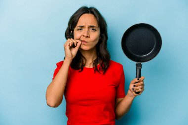 Mavi arka planda izole edilmiş bir tava tutan genç İspanyol kadın dudaklarında parmaklar ile bir sır saklıyor..