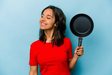 Mavi arka planda izole edilmiş bir tavayı tutan genç İspanyol kadın gülümsüyor ve keyifli görünüyor..