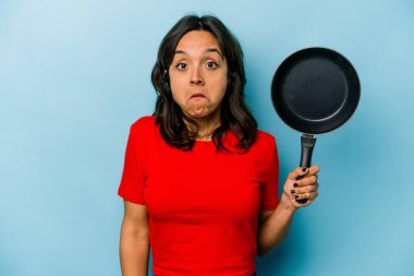 Mavi arka planda izole edilmiş bir tavayı tutan genç İspanyol kadın omuzlarını silker ve gözleri açık kalır..