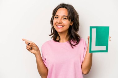 Beyaz arka planda izole edilmiş, gülümseyen ve işaret eden, boş bir alanda bir şey gösteren, acemi sürücü tabelasını tutan genç İspanyol kadın..