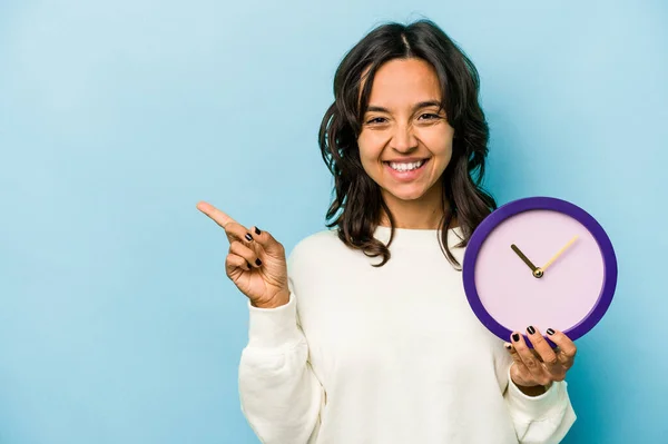 Молода Іспаномовна Жінка Тримає Годинник Ізольовано Синьому Фоні Посміхаючись Вказуючи — стокове фото