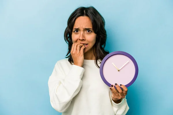 Young Hispanic Woman Holding Clock Isolated Blue Background Biting Fingernails — Stock fotografie