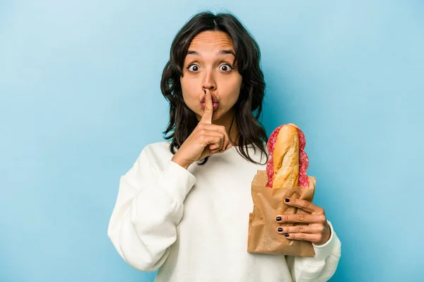 Young hispanic woman eating a sandwich isolates on blue background keeping a secret or asking for silence.