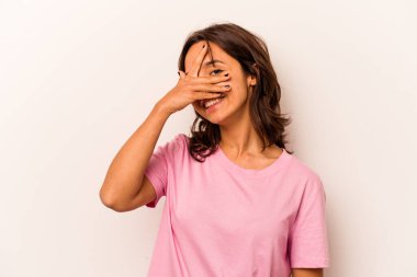 Beyaz arka planda izole edilmiş genç İspanyol kadın kameraya parmaklarıyla göz kırpıyor, yüzünü kapatmaktan utanıyor..