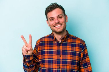 Mavi arka planda izole edilmiş genç beyaz adam zafer işareti gösteriyor ve gülümsüyor..