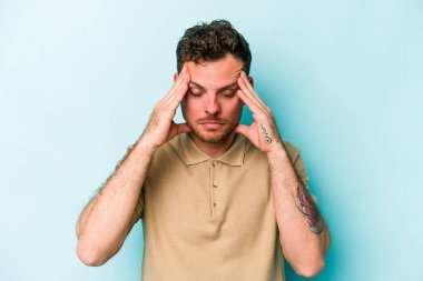 Genç beyaz adam mavi arka planda izole edilmiş tapınaklara dokunuyor ve başı ağrıyor..