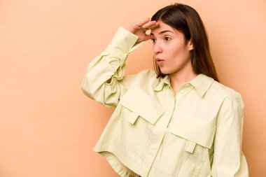 Latin kökenli genç bir kadın bej arka planda izole edilmiş, uzaklara bakıyor ve elini alnına koyuyor..