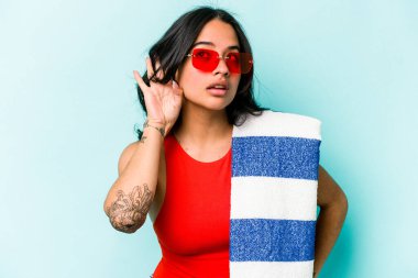 Mavi arka planda izole edilmiş plaj havlusu tutan genç İspanyol kadın bir dedikoduyu dinlemeye çalışıyor..