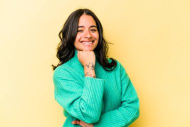 Sarı arka planda izole edilmiş genç İspanyol kadın mutlu ve kendinden emin gülümsüyor, elleriyle çenesine dokunuyor..