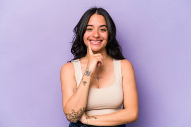 Mor arka planda izole edilmiş genç İspanyol kadın mutlu ve kendinden emin gülümsüyor, elleriyle çenesine dokunuyor..