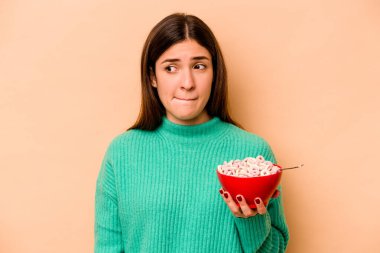 Latin kökenli genç bir kadın bej arka planda izole edilmiş mısır gevreği yiyor. Kafası karışık, kararsız ve kendinden emin değil..