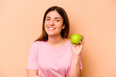 Elma tutan genç İspanyol kadın bej arka planda izole edilmiş gülüyor ve eğleniyor..