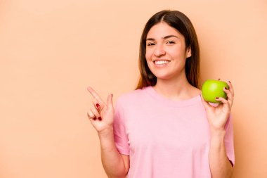 Elinde bej arka planda izole edilmiş bir elma tutan genç İspanyol kadın gülümsüyor ve boş alanda bir şey gösteriyor..