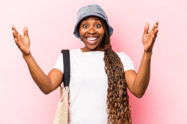 Genç Afrikalı Amerikalı kadın pembe arka planda izole bir şekilde kumsala gidiyor. Hoş bir sürpriz alıyor, heyecanlı ve el kaldırıyor..