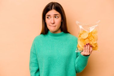 Bej arka planda izole edilmiş bir paket cipsi tutan genç İspanyol kadın kafası karışmış ve kendinden emin değil..
