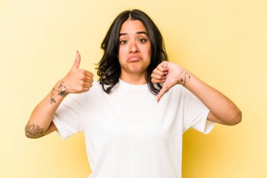 Sarı arka planda izole edilmiş genç İspanyol kadın baş parmağını kaldırıyor, zor bir seçim.