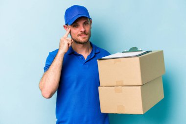 Kafkasyalı genç teslimatçı mavi arka planda izole edilmiş, parmağıyla tapınağı işaret eden, düşünen, bir göreve odaklanmış..