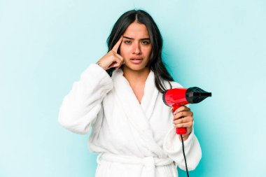 Elinde kurutma makinesi tutan genç İspanyol kadın mavi arka planda izole edilmiş, parmağıyla tapınağı işaret eden, düşünen, bir göreve odaklanmış..