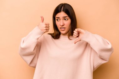 Latin kökenli genç bir kadın bej arka planda izole edilmiş. Baş parmağını kaldırmış, baş parmağını aşağı gösteriyor. Zor seçim konsepti.