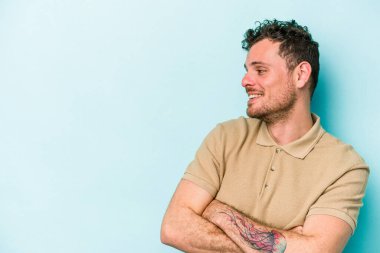 Mavi arka planda izole edilmiş genç beyaz adam kollarını kavuşturarak gülümsüyor..