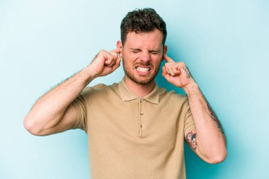Genç beyaz adam mavi arka planda izole edilmiş parmaklarıyla kulaklarını kapatıyor, gürültülü bir ortam tarafından stresli ve umutsuz..