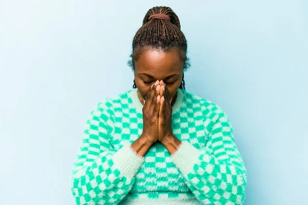 Genç Afrikalı Amerikalı bir kadın mavi arka planda izole edilmiş dua ediyor, bağlılık gösteriyor, dindar biri ilahi ilham arıyor..