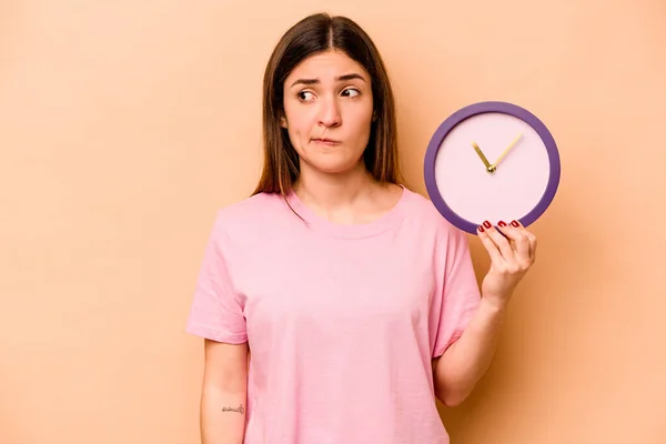 Mujer Hispana Joven Sosteniendo Reloj Aislado Sobre Fondo Beige Confundida —  Fotos de Stock