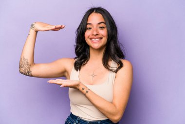 Mor arka planda izole edilmiş genç İspanyol kadın işaret parmaklarıyla küçük bir şey tutuyor, gülümsüyor ve kendinden emin..