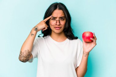 Mavi arka planda izole edilmiş bir elma tutan genç İspanyol kadın işaret parmağıyla yaptığı hayal kırıklığını gösteriyor..