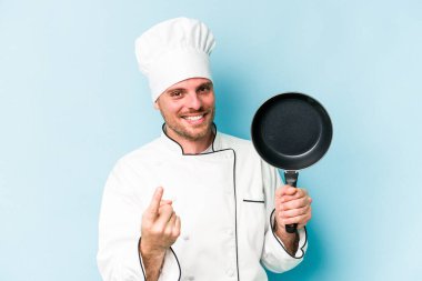 Kafkasyalı genç aşçı elinde uçan tavayla mavi arka planda izole edilmiş bir şekilde seni işaret ediyor sanki davet ediyormuş gibi..