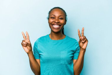 Genç Afrikalı Amerikalı kadın mavi arka planda izole edilmiş zafer işareti gösteriyor ve gülümsüyor..