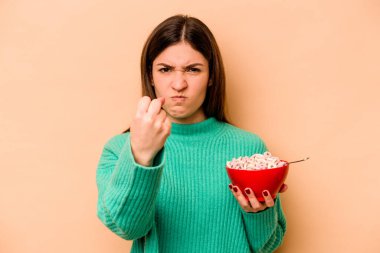 Latin kökenli genç bir kadın bej arka planda izole edilmiş mısır gevreği yiyor. Kameraya yumruk, agresif yüz ifadesi gösteriyor..