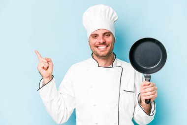 Kafkasyalı genç aşçı elinde uçan bir tavayla mavi arka planda izole edilmiş gülümsüyor ve boş alanda bir şey gösteriyor..