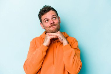 Mavi arka planda izole edilmiş genç beyaz adam ellerini çenesinin altında tutuyor, mutlu bir şekilde yana bakıyor..