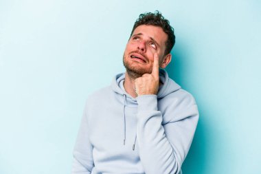 Kafkasyalı genç adam mavi arka planda izole edilmiş, ağlıyor, bir şeyden memnun değil, acı ve kafa karışıklığı konsepti..