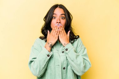 Sarı arka planda izole edilmiş genç İspanyol kadın şok olmuş, ağzı ellerle kaplanmış, yeni bir şey keşfetmeye hevesli..