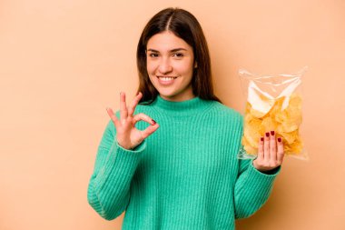 Latin kökenli genç bir kadın elinde bir paket cips tutuyor. Bej arka planda izole edilmiş. Neşeli ve kendinden emin..